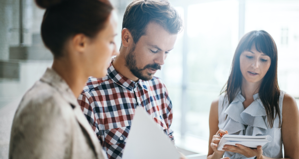 This image showcases 3 individuals, one young man and 2 young women, all standing and having a discussion. They are holding paper documents in their hands
