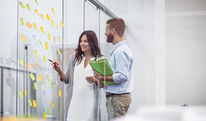 A man and lady looking at a glass wall with sticky notes on it. The lady is pointing to a sticky note and the man is holding a folder.